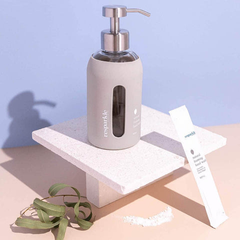 Soap dispenser with a pump and a sachet of foaming hand wash on a light pink surface with a purple background.