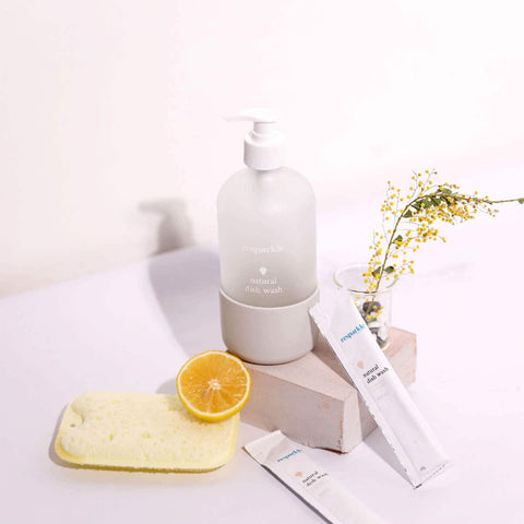 Dishwash kit products including a bottle, sachet, and sponge on a white surface with a lemon and flowers.