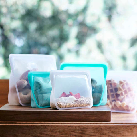 Collection of reusable stasher stand up bags in clear and aqua colours, each filled with food and sitting on a wooden board. 