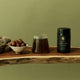 Jar of coffee, glass of coffee, and bowl of figs on a wooden surface with a green background