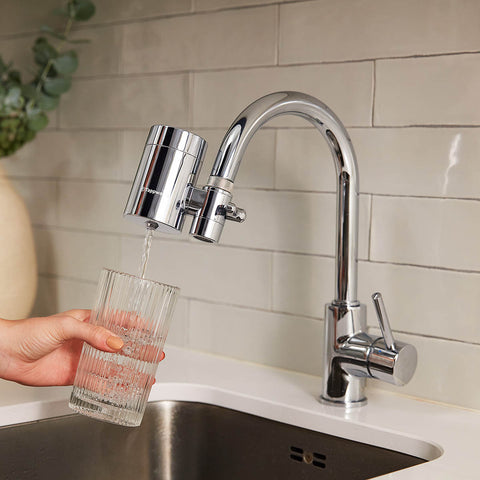 Person filling a glass with water from a kitchen faucet with a water filter attachment.
