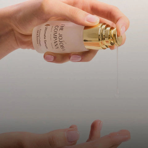 Hand holding a bottle of 'The Jojoba Company' serum with a gold cap, dripping serum onto skin.