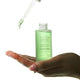 Hand holding a green bottle with a dropper labeled 'The Jojoba Company Jojoba Boost Skin Renewal' on a white background.