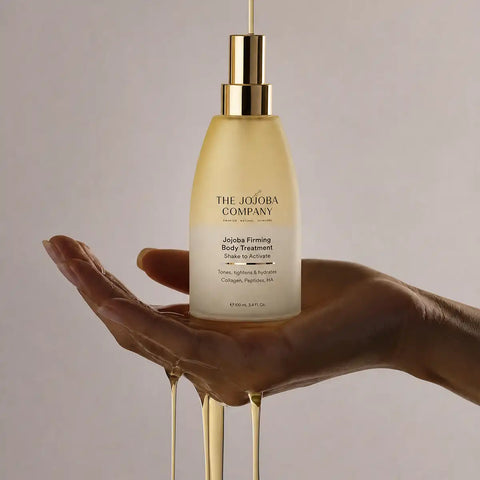 A hand holds a frosted bottle of The Jojoba Company Jojoba Firming Body Treatment, with golden honey-like product dripping from the bottle and around the hand against a soft beige background.