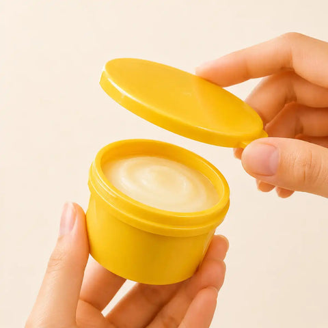 A small round yellow plastic jar with its lid off, held in two hands against a plain light beige background. The jar contains a pale cream-colored balm or cream with a swirled texture.