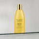A bottle of The Jojoba Company Jojoba Luxe Shower Oil, filled with golden yellow liquid and topped with a metallic gold cap, resting on a glass shelf against a textured, rain-slicked gray background.
