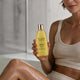 A woman in a beige ribbed tank top holds a bottle of The Jojoba Company Jojoba Shower Oil. The bottle is a translucent golden yellow with a shiny gold cap.