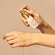 Hand holding a bottle of The Jojoba Company Pigmentation Oil product with a swatch on skin against a beige background.