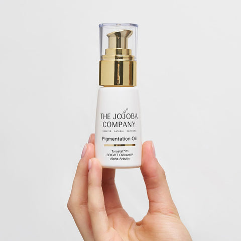 Hand holding a bottle of The Jojoba Company Pigmentation Oil against a light gray background.