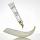 A white tube of The Jojoba Company Ultimate Plumping Lip Treatment with a gold cap, shown dispensing a drop of cream onto a curved white dish against a clean white background.