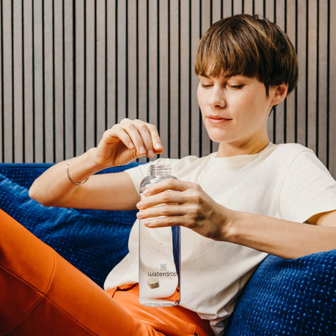 Person holding a water bottle with a brand logo, sitting on a blue couch.