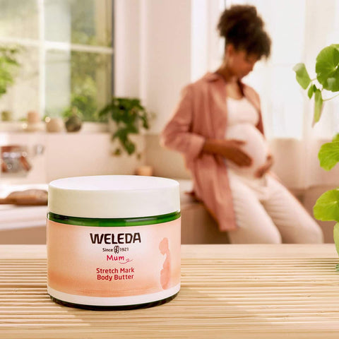 Weleda Stretch Mark Body Butter 150ml jar on a table with a blurred background of a woman sitting on a couch.
